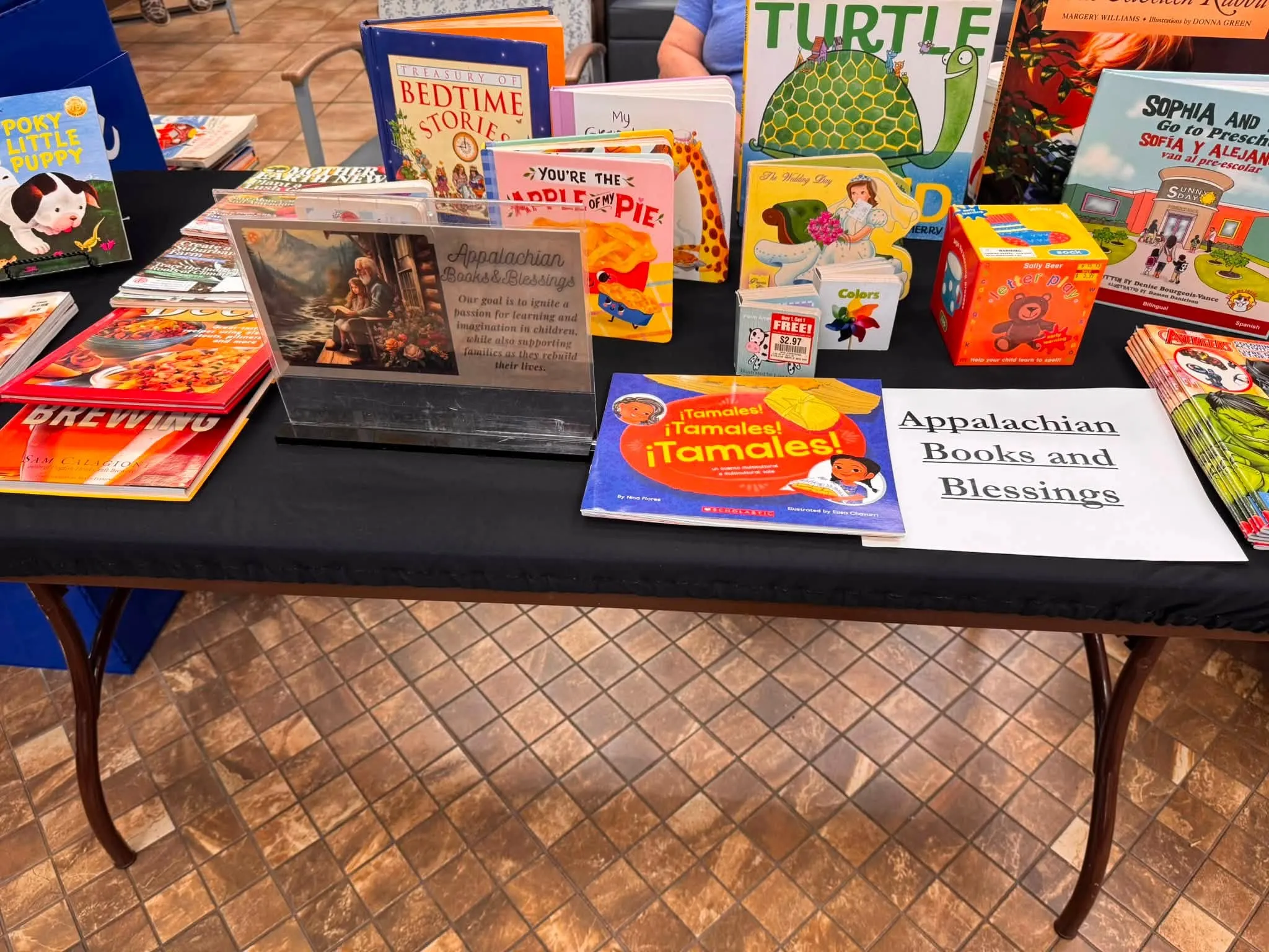 A table draped in black cloth, covered with children's picture books, board books, and magazines, beside an Appalachian Books & Blessings sign.
